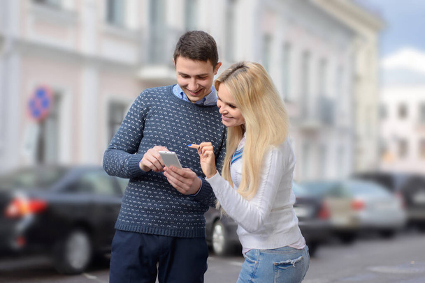 κορίτσι ξανθιά και ο τύπος ψάχνει κάτι στο κινητό τηλέφωνο και χαμογελώντας στο δρόμο της πόλης - Φωτογραφία, εικόνα