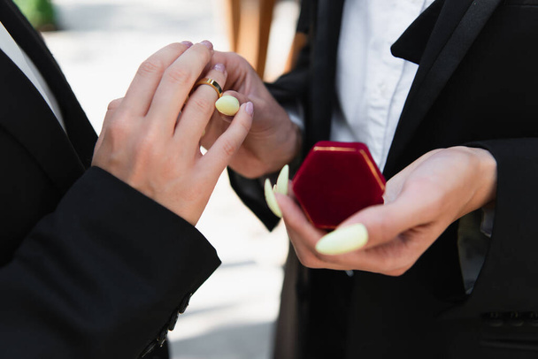 Cropped view of lesbian woman in suit wearing wedding ring on finger of girlfriend outdoors  - Фото, изображение