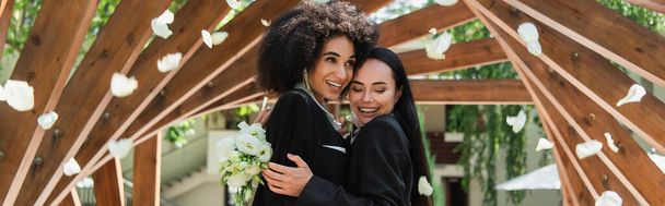Lesbian african american woman hugging cheerful girlfriend with wedding bouquet under falling petals in park, banner  - Фото, изображение