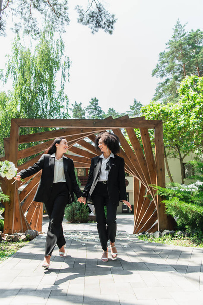 Smiling interracial lesbian couple with wedding bouquet holding hands in park  - 写真・画像