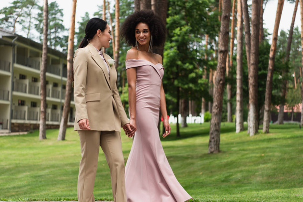 笑顔異人種間のレズビアンカップルでドレスとスーツ保持手で公園を歩く  - 写真・画像