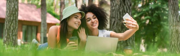 Smiling interracial lesbian couple with coffee taking selfie near laptop in park, banner  - Foto, Bild