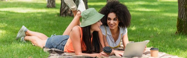 Smiling interracial lesbian couple using devices near paper cups on blanket in park, banner  - Photo, Image
