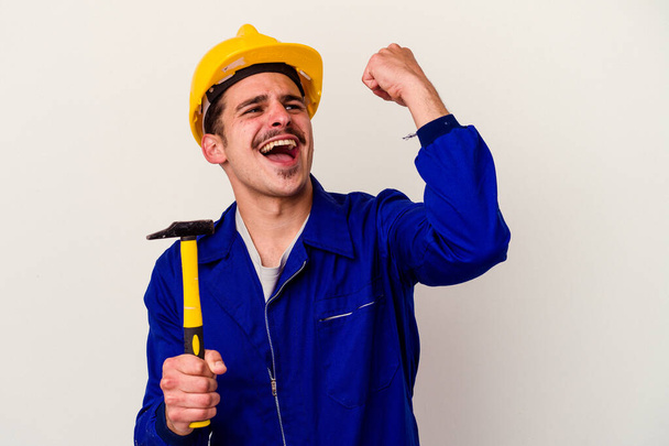 Молодий кавказький працівник тримає молоток ізольований на білому тлі, піднімаючи кулак після перемоги, концепція переможця
. - Фото, зображення