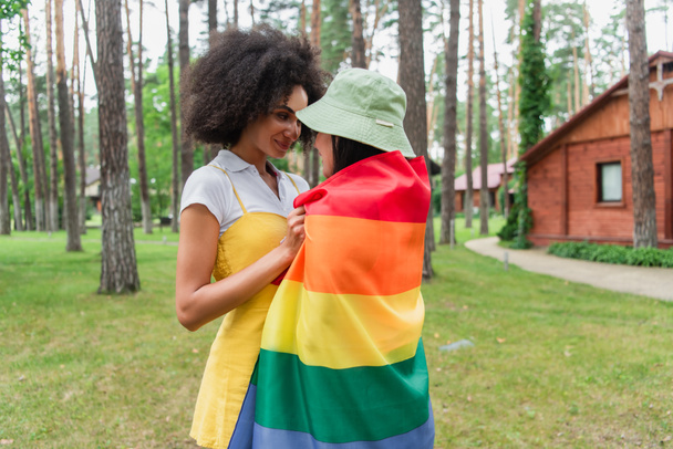 Smiling african american woman hugging girlfriend in lgbt flag outdoors  - Foto, immagini