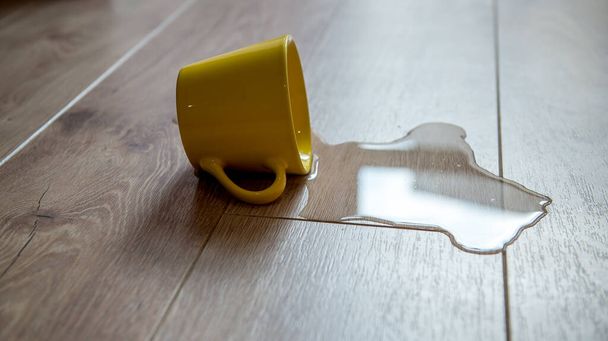 Yellow cup of water spilled on a wooden laminate floor. Moisture protection of a floor. Waterproof parquet board with spilled water. - Photo, Image