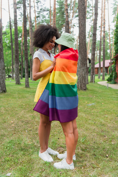 Positive african american woman hugging girlfriend in lgbt flag in park  - Photo, image