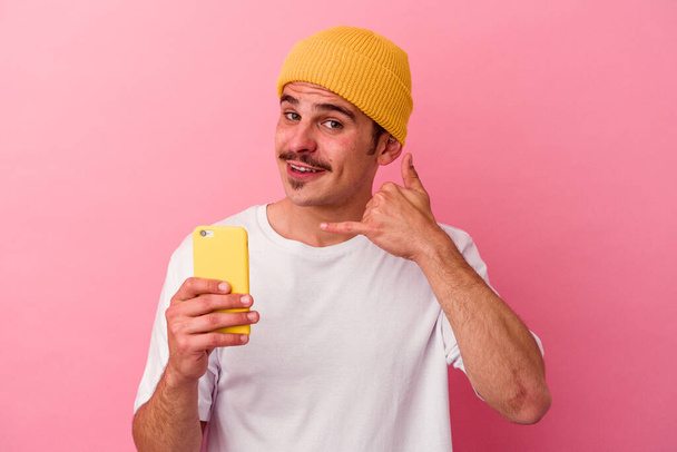 Junger kaukasischer Mann mit einem Mobiltelefon auf rosa Hintergrund, das eine Handyanruf-Geste mit den Fingern zeigt. - Foto, Bild