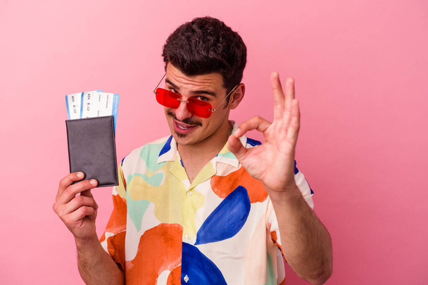 Νεαρός ταξιδιώτης καυκάσιος άνδρας που κατέχει διαβατήριο απομονωμένος σε ροζ φόντο χαρούμενος και σίγουρος που δείχνει εντάξει χειρονομία. - Φωτογραφία, εικόνα