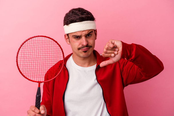 Young caucasian man playing badminton isolated on pink background showing a dislike gesture, thumbs down. Disagreement concept. - Foto, Imagen