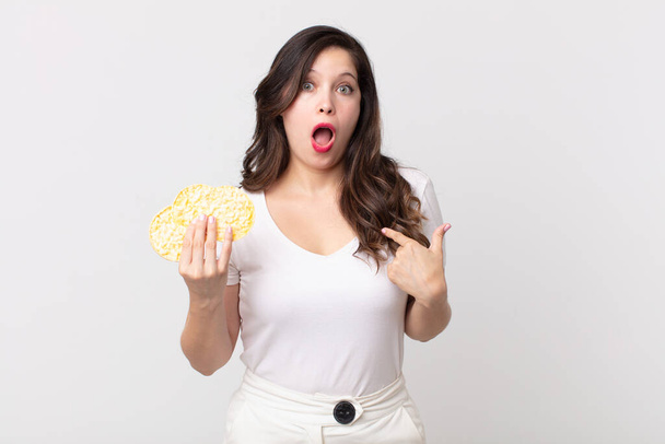 pretty woman looking shocked and surprised with mouth wide open, pointing to self and holding a diet rice cakes - Foto, Imagen