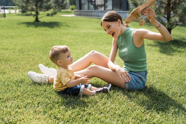 счастливая молодая мать, держащая игрушечный биплан и глядя на веселого малыша сына на траве - Фото, изображение