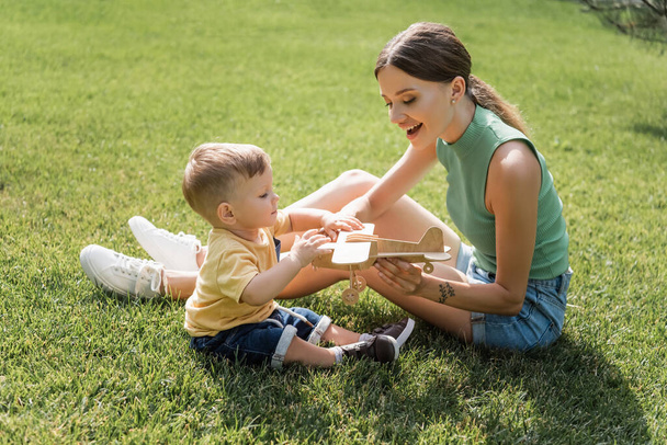 cheerful mother holding toy biplane near toddler son on grass - Fotografie, Obrázek