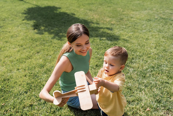 smiling mother holding toy biplane near toddler son on grass - Foto, Imagen
