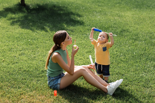 young mother sitting on grass and blowing soap bubbles near toddler son  - Foto, Bild
