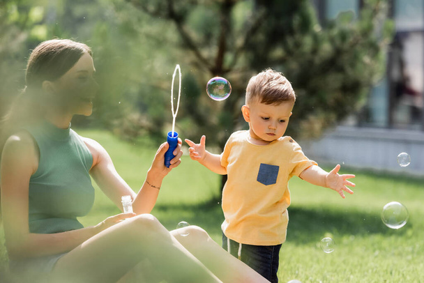 blurred and joyful mother looking at toddler son near soap bubbles  - Foto, Imagem