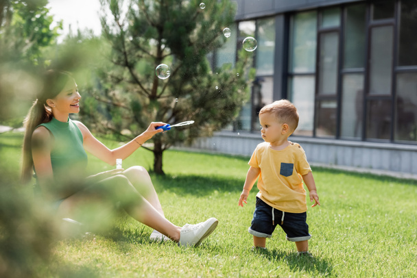 joyful mother looking at toddler son near soap bubbles  - Foto, afbeelding