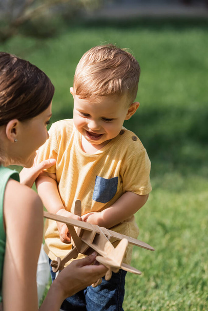 blurred cheerful mother near smiling toddler boy with wooden biplane  - Photo, Image