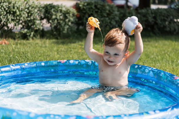 χαρούμενο αγοράκι που παίζει με παιχνίδια από καουτσούκ και ρίχνει νερό σε φουσκωτή πισίνα έξω  - Φωτογραφία, εικόνα
