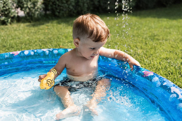 niño pequeño sentado en piscina inflable con juguete de goma afuera - Foto, imagen