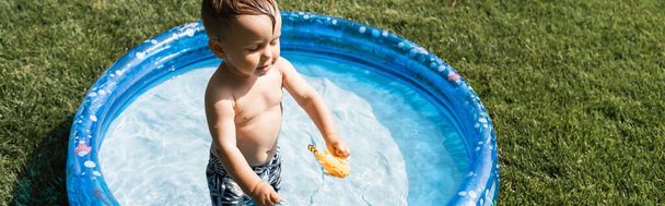 high angle view of cheerful toddler boy standing in inflatable pool with rubber toy, banner - Foto, immagini