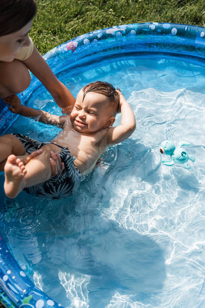 blurred mother supporting toddler son with closed eyes swimming in inflatable pool - Photo, Image