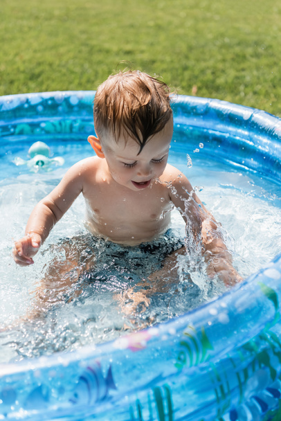 νήπιο αγόρι κάθεται σε φουσκωτή πισίνα και κάνει πιτσιλιές νερού  - Φωτογραφία, εικόνα