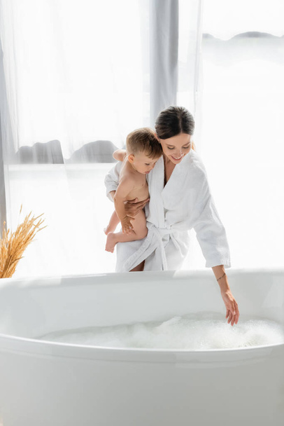 caring mother in bathrobe holding in arms naked toddler son near bathtub with bath foam  - Zdjęcie, obraz