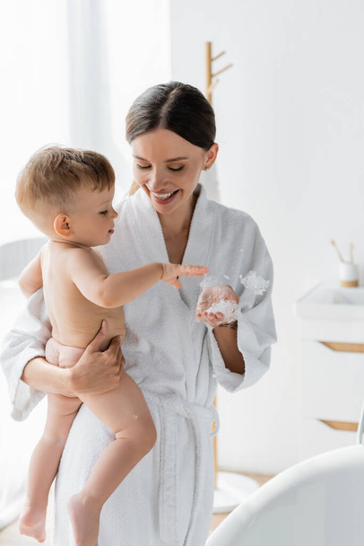 cheerful mother in bathrobe holding in arms toddler son near bathtub with bath foam  - Zdjęcie, obraz