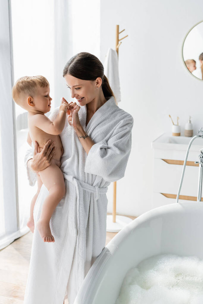 cheerful mother in bathrobe holding in arms and touching fingers with toddler son near bathtub with bath foam  - Zdjęcie, obraz