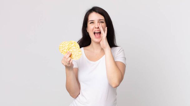 pretty woman feeling happy,giving a big shout out with hands next to mouth and holding a diet rice cakes - Fotoğraf, Görsel
