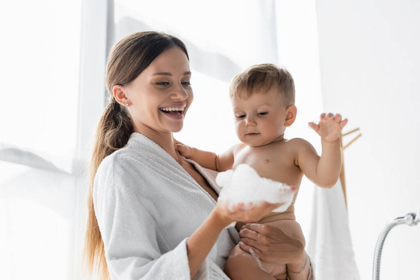 smiling mother in bathrobe holding bath foam near naked toddler son  - 写真・画像