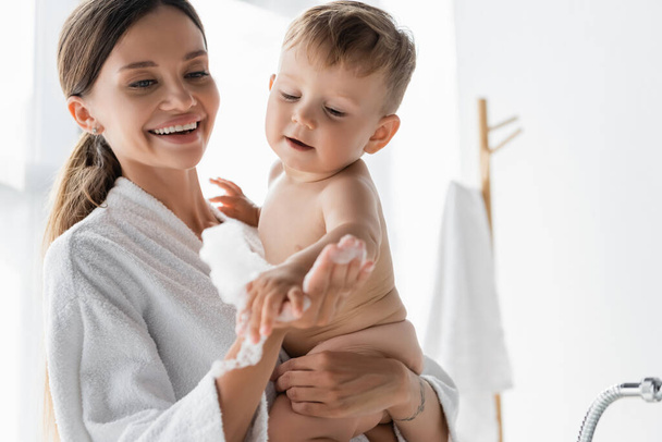 smiling mother in bathrobe holding bath foam and naked toddler son in bathroom - Фото, изображение