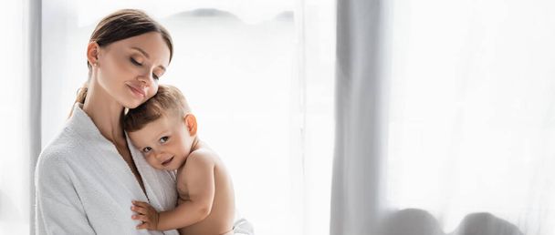 ευχαριστημένη μητέρα με μπουρνούζι που κρατά στην αγκαλιά της γυμνό γιο νήπιο, πανό - Φωτογραφία, εικόνα