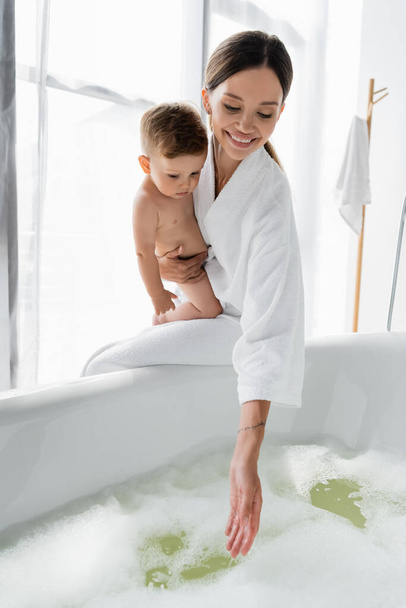 pleased and tattooed mother in bathrobe holding in arms nude toddler son and reaching water in bathtub  - Foto, afbeelding