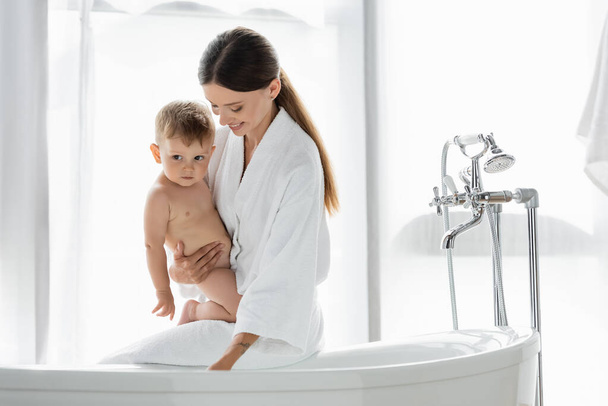 pleased woman in bathrobe holding in arms toddler son near bathtub  - Photo, Image