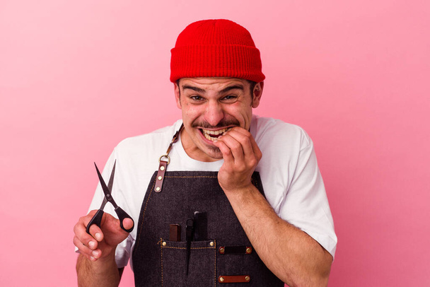 Молодой кавказский парикмахер держит ножницы изолированные на розовом фоне кусая ногти, нервный и очень тревожный. - Фото, изображение