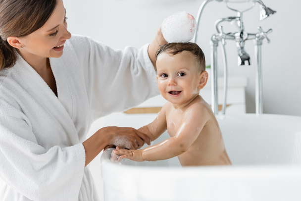 cheerful mother in bathrobe bathing happy toddler son in bathtub  - Foto, Imagem