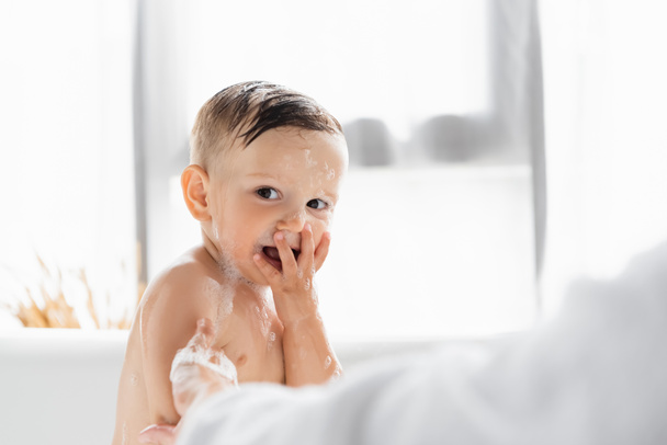 blurred mother in bathrobe bathing wet and happy toddler son in bathtub  - Foto, afbeelding