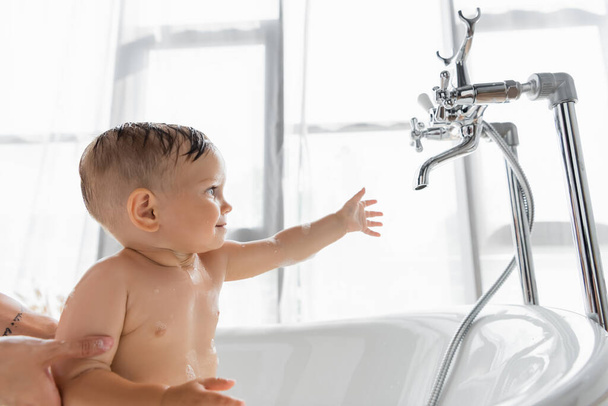 tattooed mother holding toddler son reaching faucet while bathing in bathtub  - Photo, image