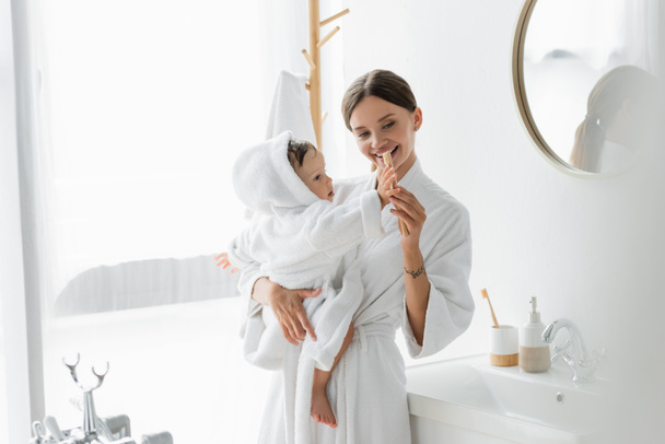 madre feliz sosteniendo en brazos hijo pequeño en albornoz y cepillo de dientes - Foto, imagen