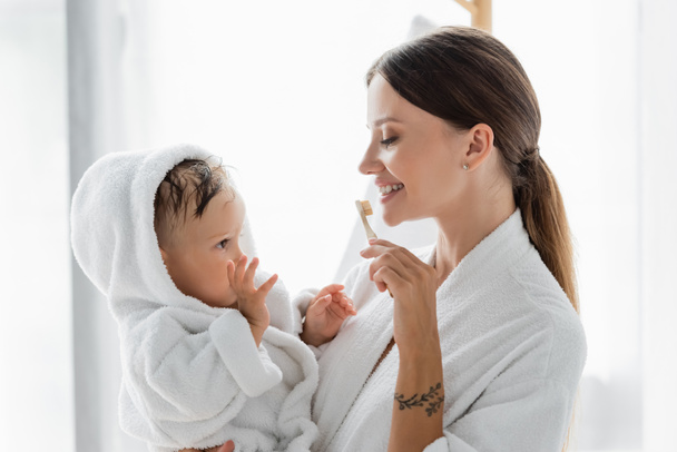 smiling mother brushing teeth and holding in arms toddler son in bathrobe  - Foto, immagini