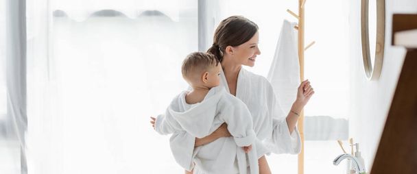 πλαϊνή άποψη του χαρούμενα μητέρα εκμετάλλευση στην αγκαλιά παιδί γιο και κοιτάζοντας καθρέφτη στο μπάνιο, πανό - Φωτογραφία, εικόνα