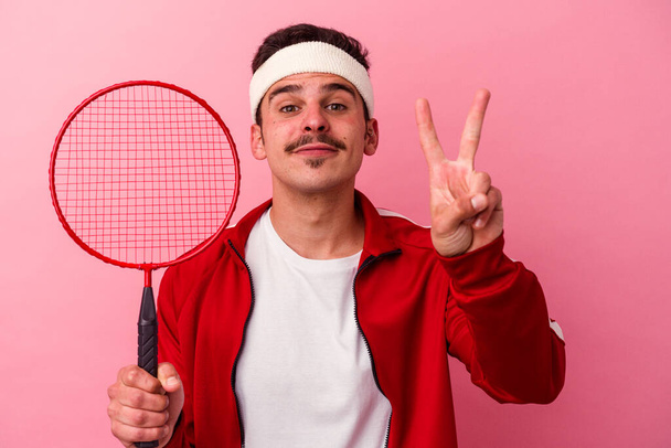 Mladý běloch hraje badminton izolované na růžovém pozadí ukazující číslo dvě s prsty. - Fotografie, Obrázek