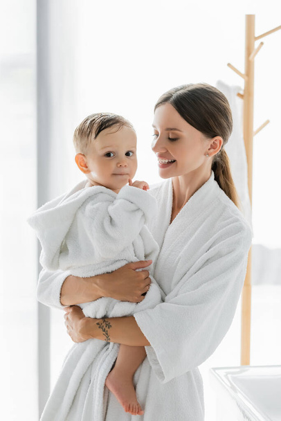 happy mother looking at toddler son in bathrobe - Foto, Imagen