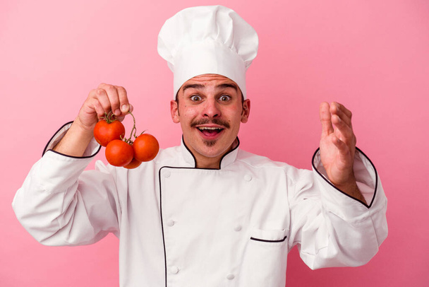 Jonge Kaukasische chef-kok houdt tomaten geïsoleerd op roze achtergrond ontvangen van een aangename verrassing, opgewonden en het verhogen van de handen. - Foto, afbeelding