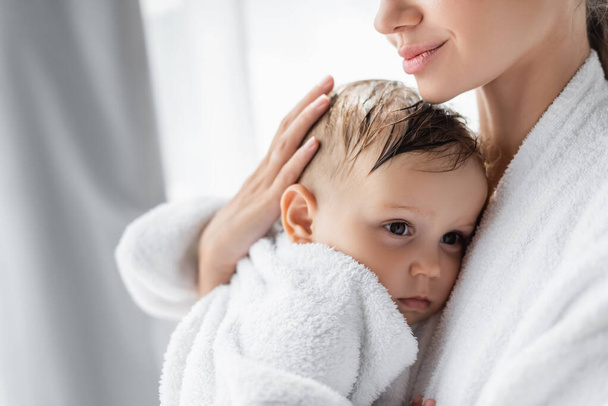 pleased young mother holding in arms toddler son in bathrobe - Photo, Image