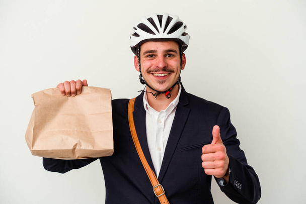 Jonge zakelijke blanke man dragen fietshelm en bedrijf nemen plaats voedsel geïsoleerd op witte achtergrond glimlachen en het verhogen van duim omhoog - Foto, afbeelding