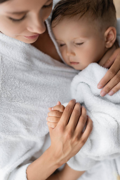 wazig en zorgzame moeder knuffelen peuter zoon met gesloten ogen in badjas - Foto, afbeelding