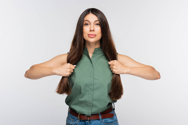 νεαρή γυναίκα προσαρμογή λαμπερά μαλλιά και κοιτάζοντας κάμερα απομονωμένη σε γκρι - Φωτογραφία, εικόνα